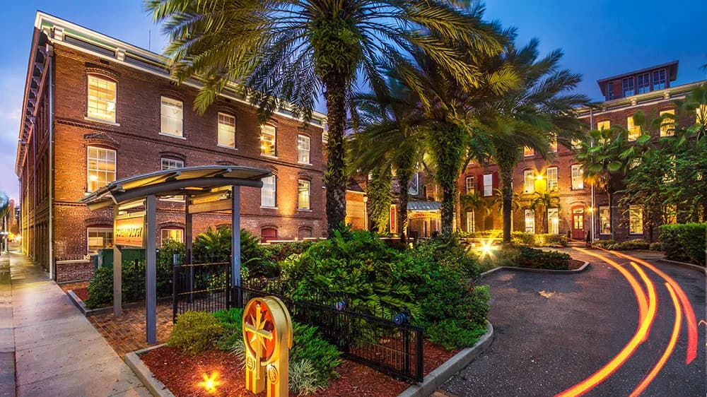 Historic four-story red brick building with palm trees and warm interior lighting during dusk, featuring a covered entryway and manicured landscaping.