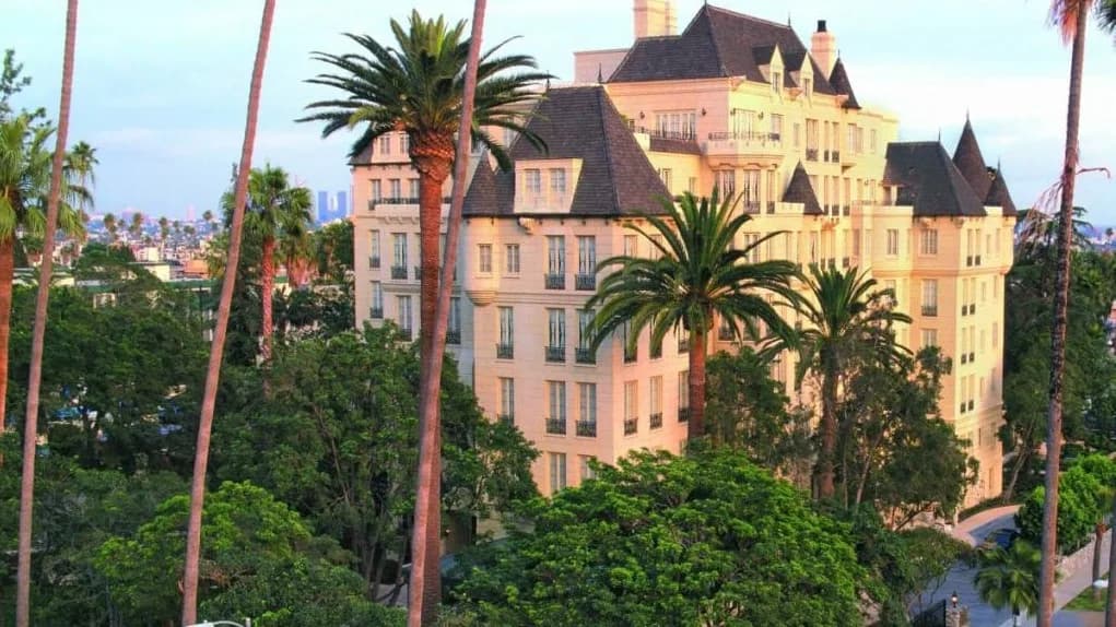 Six-story historic mansion with ornate French château architecture, mansard roof with decorative dormers, peach-colored stucco facade with balconies, surrounded by tall palm trees.