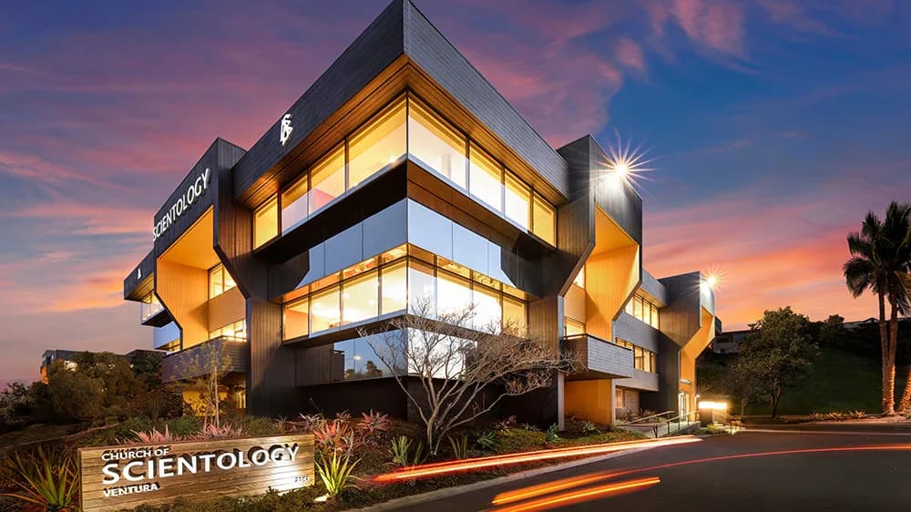 Angular contemporary building with dramatic offset geometric levels, dark metal cladding, illuminated golden interior visible through expansive windows, palm trees, and vibrant dusk sky.