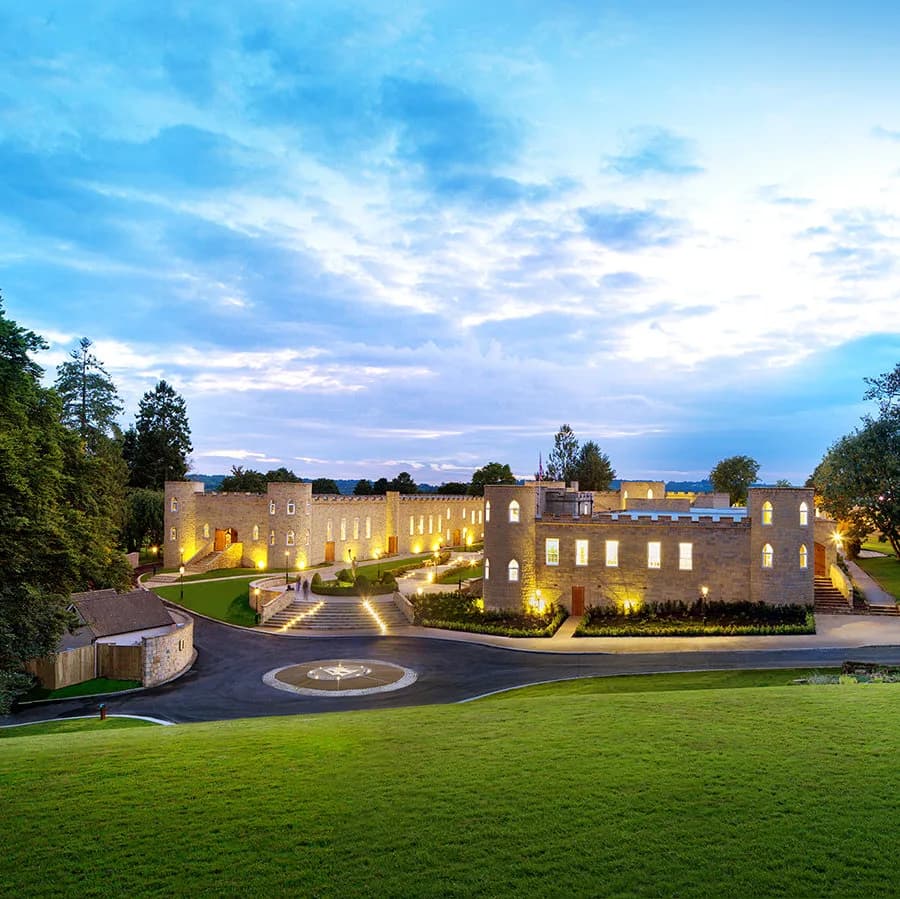 Historic stone structure with crenellated fortress-like walls and multiple stories, surrounded by manicured lawn and mature trees, illuminated at dusk with golden interior lighting.
