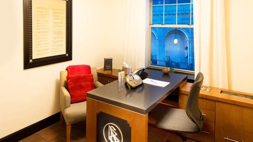 A small auditing room: a wooden desk with a chair on either side, a framed copy of the Auditor’s Code on the wall, and daylight through a curtained window.