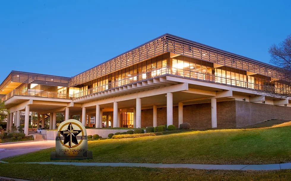 Modern multi-story building with distinctive horizontal slat roofing and dramatic cantilevers, illuminated warm interior windows, manicured grounds, and evening lighting beneath clear sky.