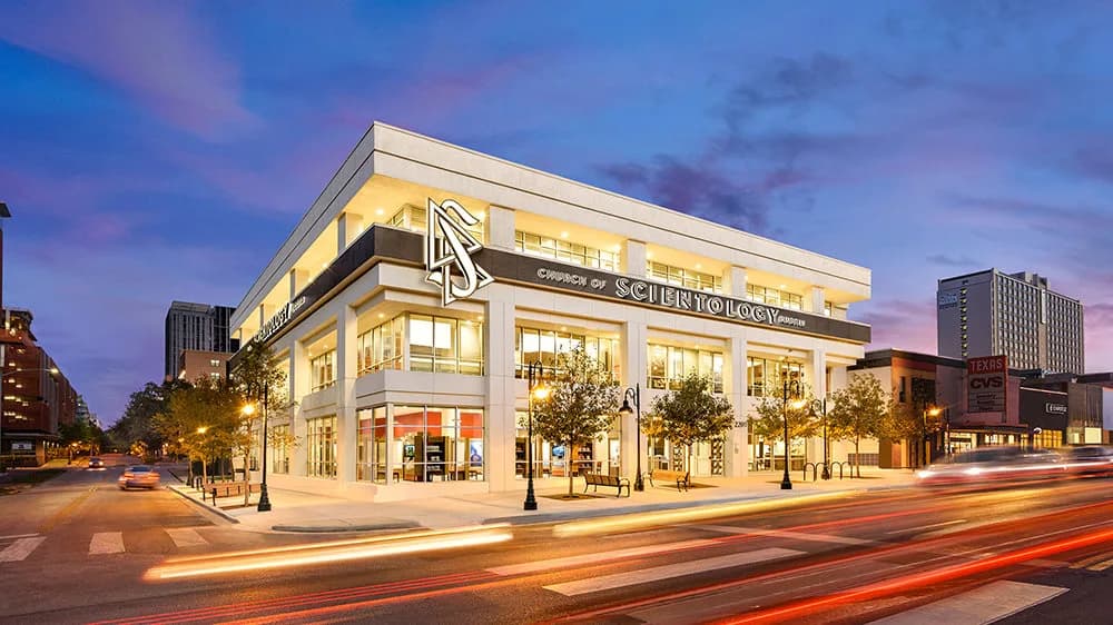 Modern white stone building with floor-to-ceiling glass windows and clean geometric lines, situated in a downtown urban setting among tall surrounding buildings, photographed at twilight.