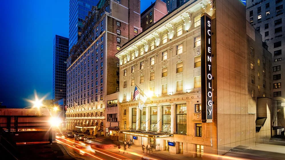 Historic multi-story building with cream-colored classical stone facade and ornamental details, featuring tall vertical SCIENTOLOGY signage, positioned on an urban street with surrounding modern buildings.