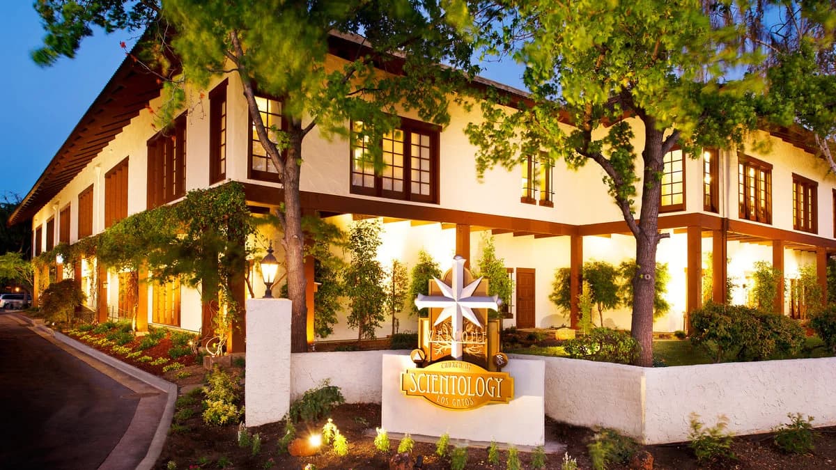 The Church of Scientology Los Gatos, a two-storey mission-style building with wooden balconies and mature trees, photographed from its driveway at dusk.